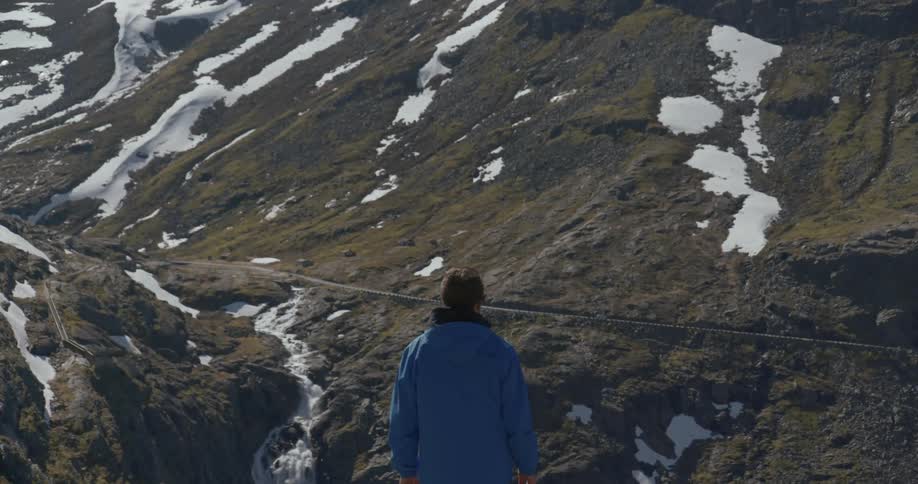 A man standing on a mountainside watching a vehicle drive along a road far below