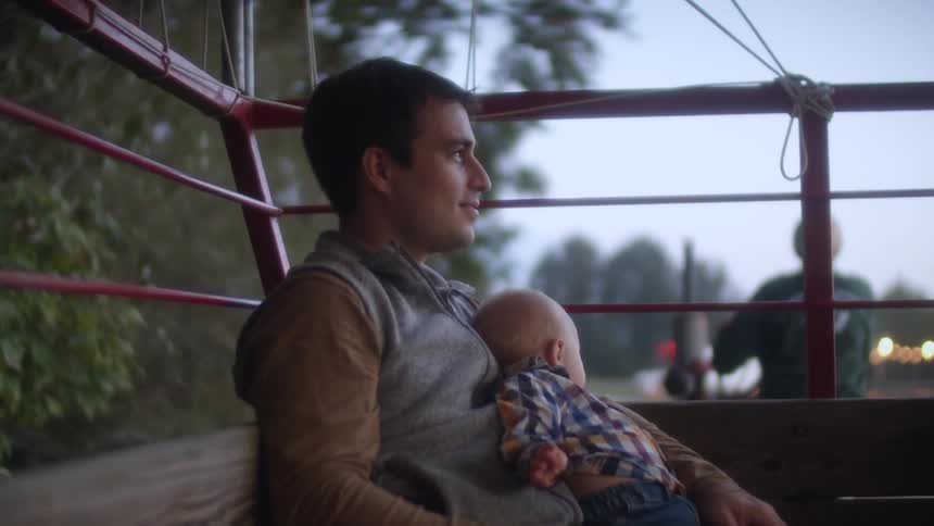 A man sitting on a tractor holding a baby with trees the sky and clouds in the background
