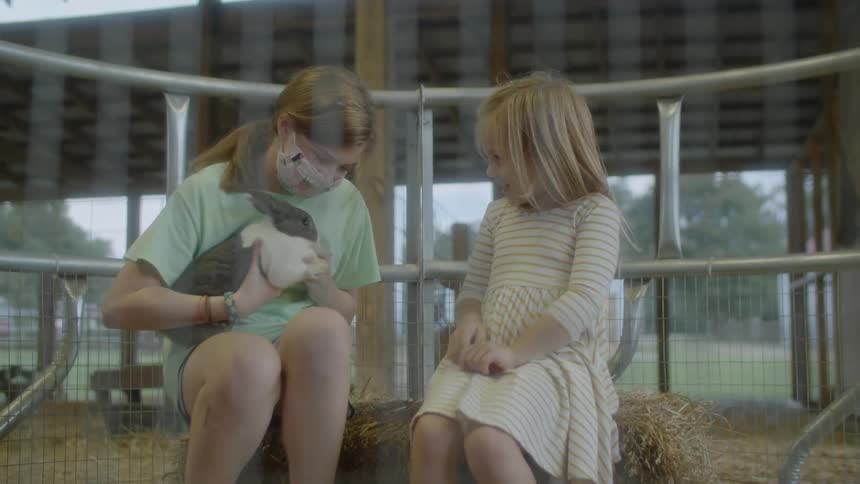 Two girls are sitting on hay in a fenced in area outdoors while they pet a bunny