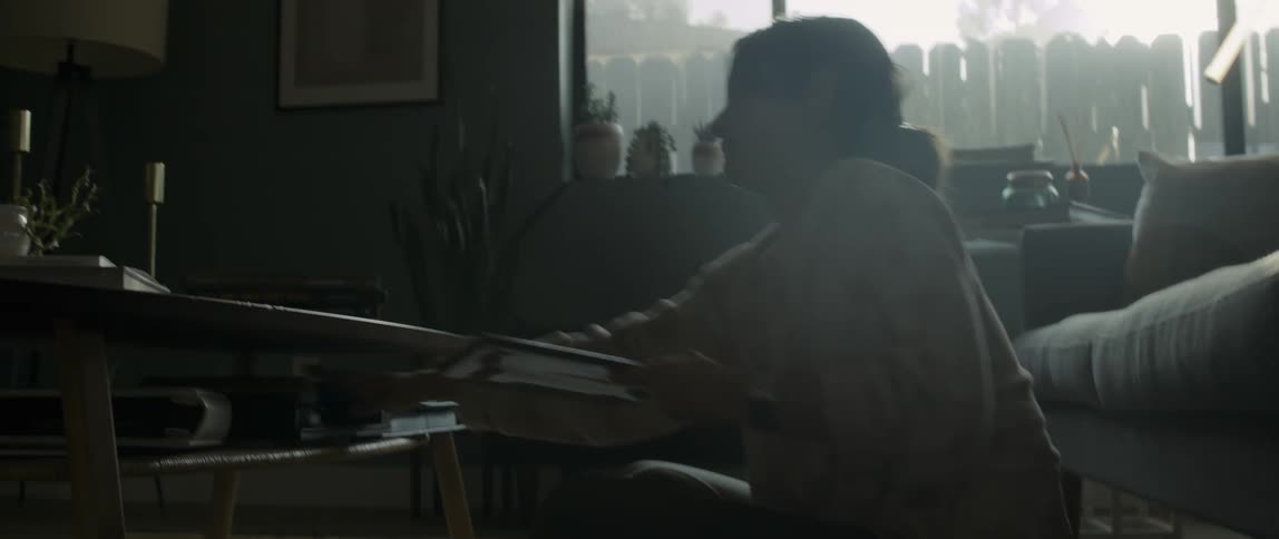 A woman sits on the floor in a living room and reaches over for a book from a table