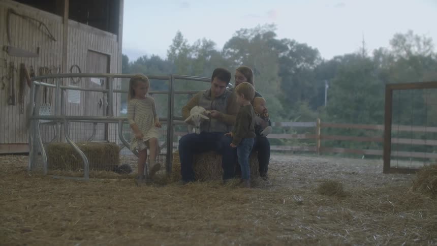 A man sitting on a bale of hay next to a woman shows the lamb he is holding to the small children standing around him