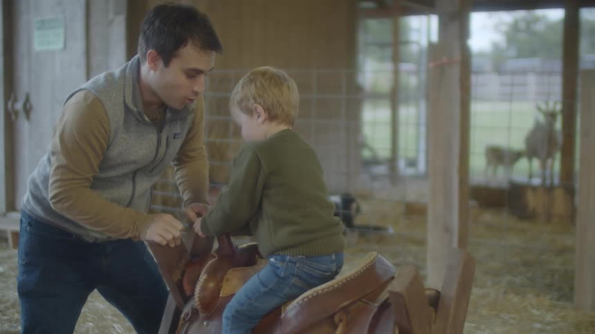 A man and a boy playing on a wooden horse with a saddle in a barn