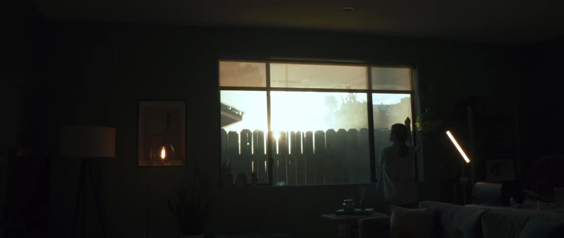 A woman lift the blinds on a window in a dark room at home at sunset