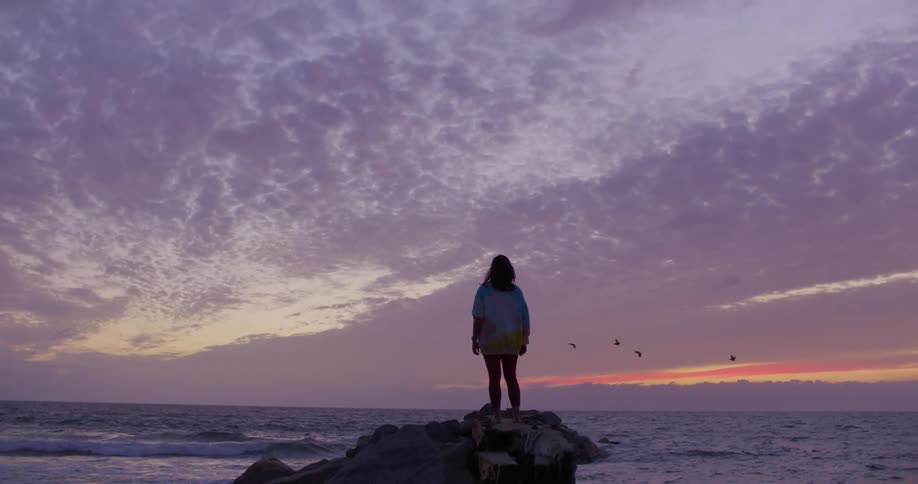 A woman is standing by the coast of an ocean at dusk