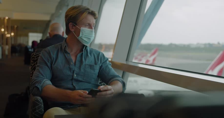 A man wearing a denim shirt and khaki pants sits in an airport looking out the window at planes as he hold a cell phone in his hands