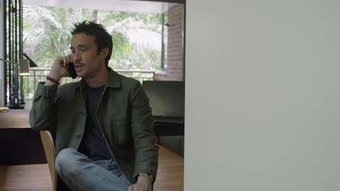 A man sits inside a home talking on the phone with a desk and laptop behind him
