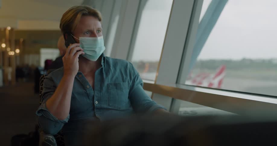 A wearing a medical mask talks on a smartphone while sitting near a large window at the airport during the day