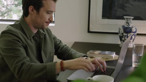 A man sitting at a desk drinks from a cup of coffee before typing on his laptop