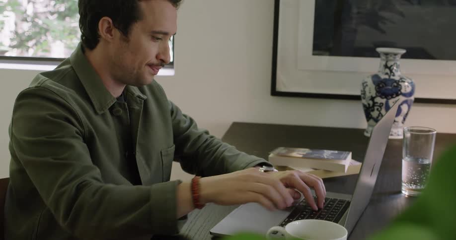A man sitting at a desk drinks from a cup of coffee before typing on his laptop