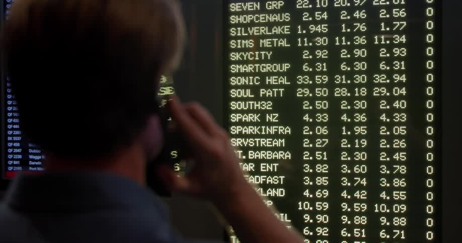 A stockbroker talks on his smartphone while looking at the names of businesses and data on multiple monitors