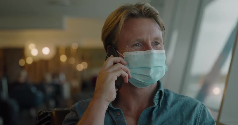 A blonde man sits indoors wearing a face mask while talking on a cell phone