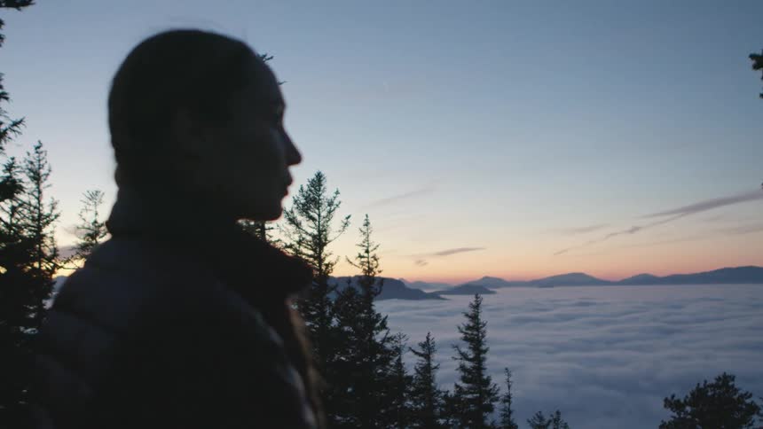 A young woman in a coat looks out at the horizon while standing high in the mountains at sunset