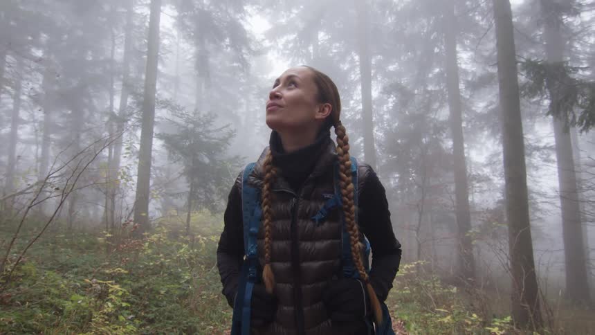 A woman leisurely looks around her surroundings while hiking through a foggy woodland during the day