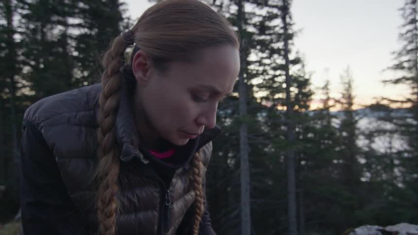 A white woman sits outdoors camping in the forest cooking and warming her hands