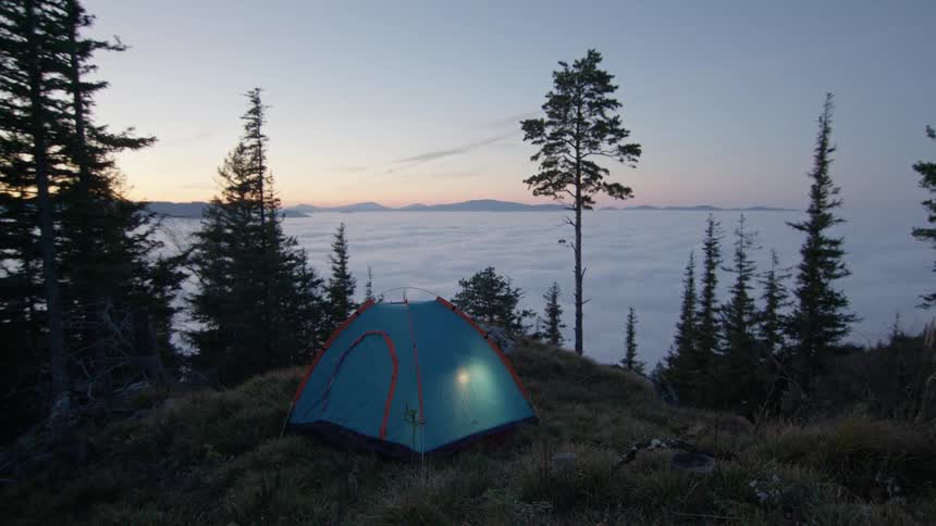 Lights shine within a tent atop a hill in the wilderness at sunset