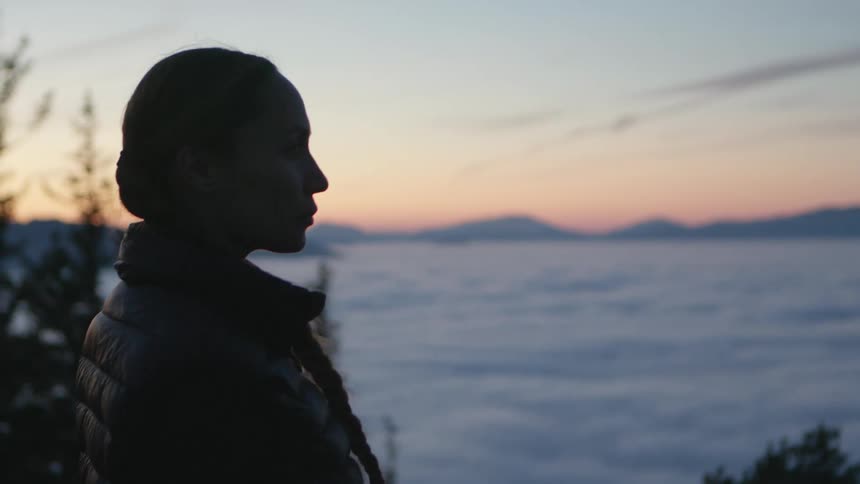 A woman standing in the woods with trees a mountain range the sky and clouds in the background