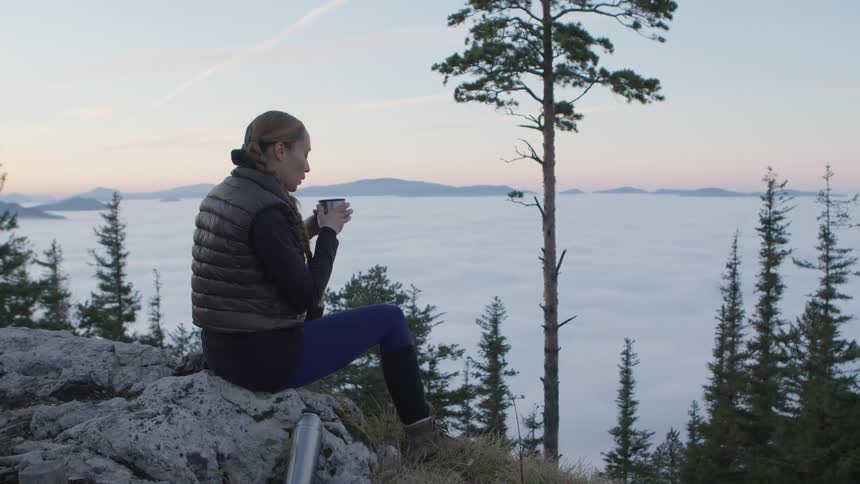 A woman sitting on a rock with a drink overlooking a lake