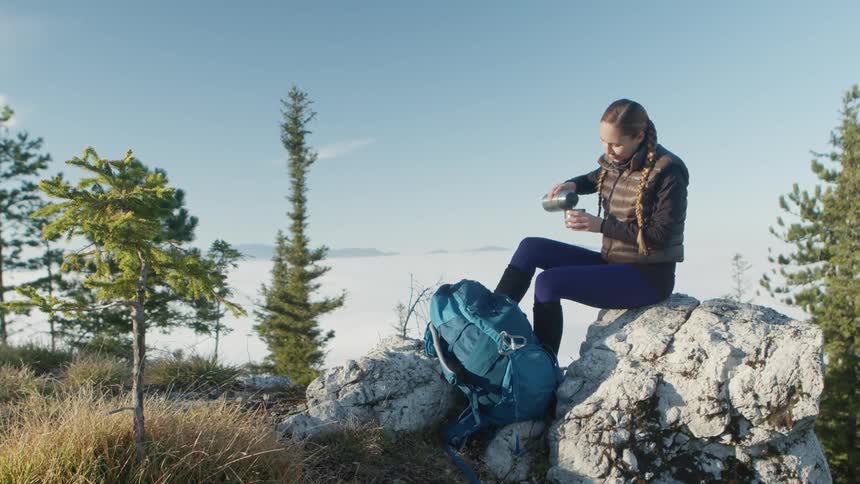 A woman sits outside in nature on a rock pouring a drink from a thermos