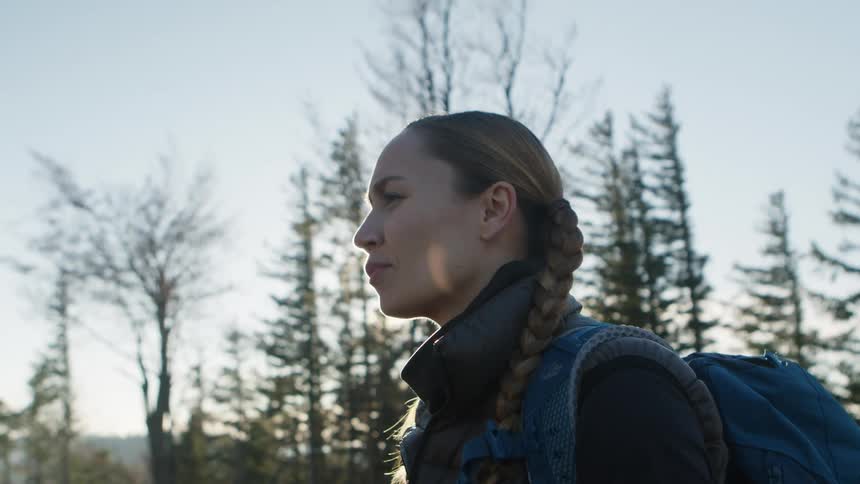 A woman with a backpack standing in the forest with trees the sky and clouds in the background
