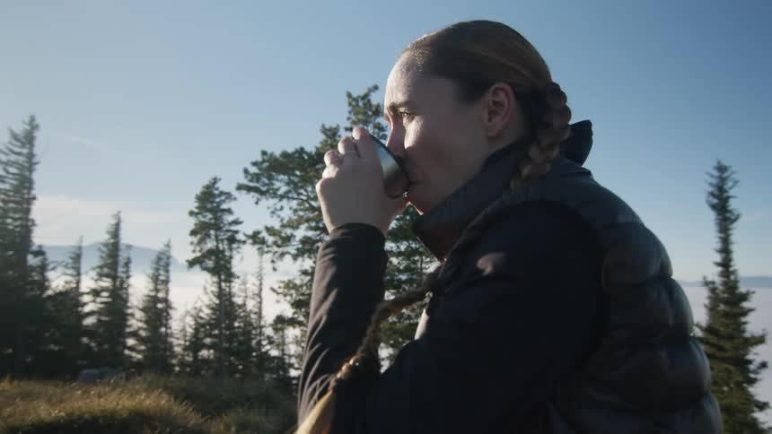 A woman in the woods drinking from a cup with trees the sky and clouds in the background