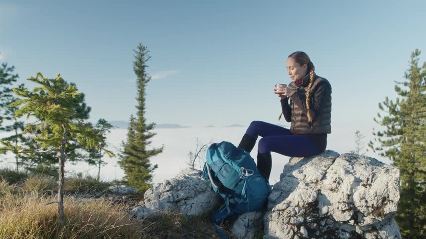 A woman takes a break from hiking to sit on a boulder and enjoy a drink