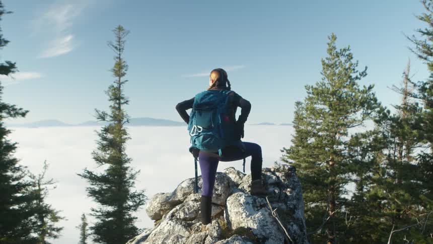 Young fit woman wearing a backpack stands atop the rocks of a mountain peak