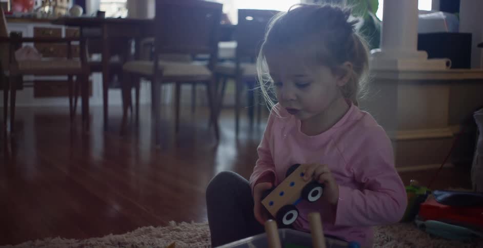 A little girl wearing a pink shirt and a pony tail sits on the floor playing with a wooden truck