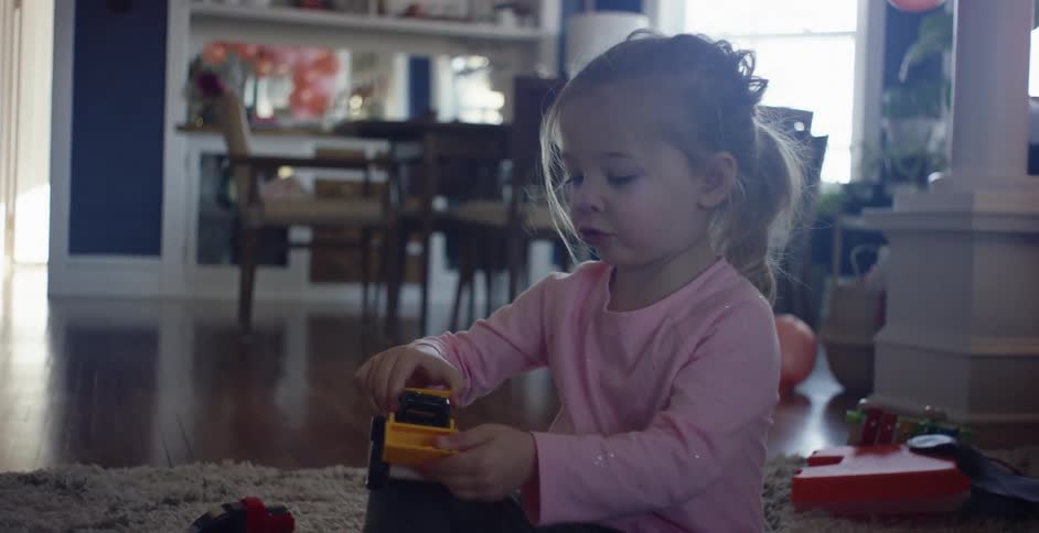 A young girl in a pink shirt sits on the floor playing with a truck in a family room