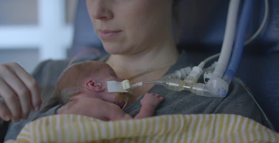 Mother holds newborn with a breathing tube while sitting in a hospital room