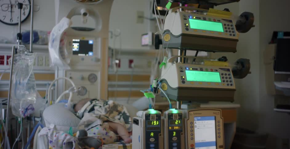 An infant sleeps in a small hospital bed surrounded by medical equipment