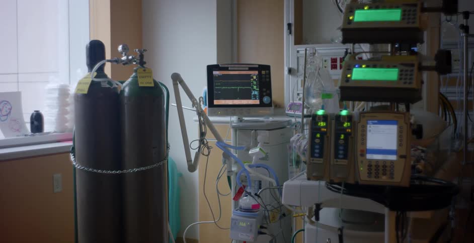 A hospital room with oxygen tanks monitors and machines with a window in the background