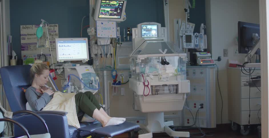 Young woman with newborn baby on her lap sitting in a hospital surrounded by medical equipment