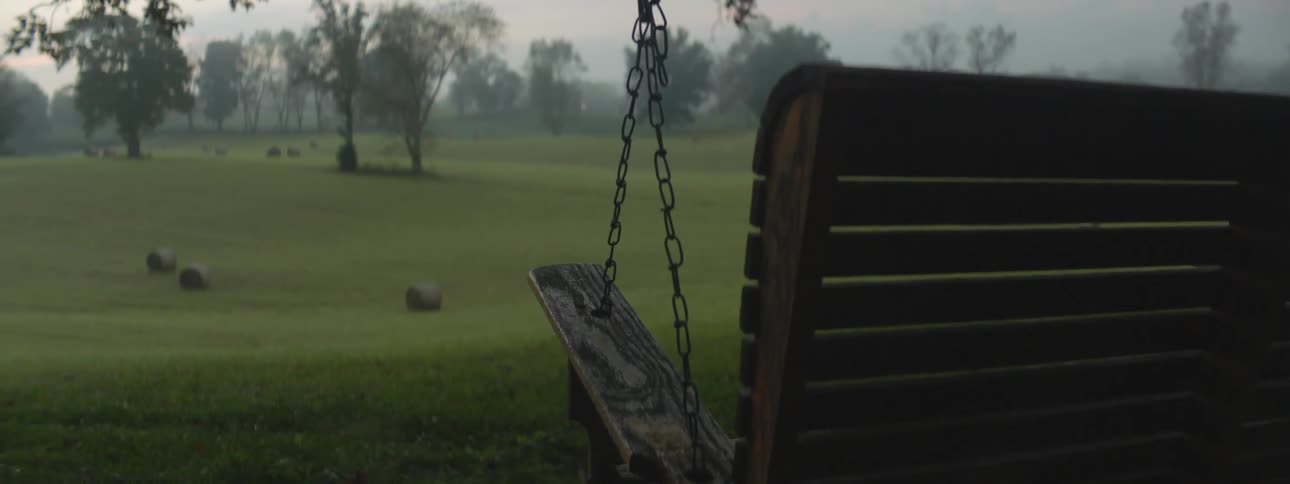 Empty porch swing sways in the wind facing an open field with hay and trees at sunrise