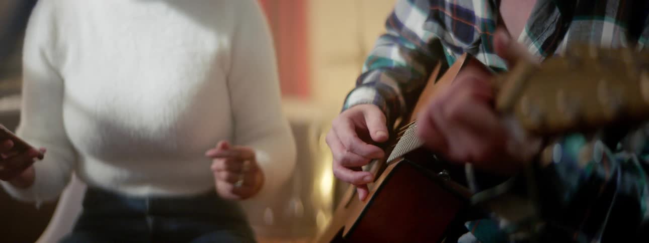 A man wearing a plaid shirt plays a guitar sitting next to a woman in a white sweater who is snapping her fingers