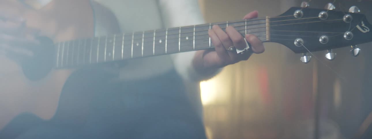 A woman wearing a white sweater sits in front of a microphone playing a guitar with smoke in the air