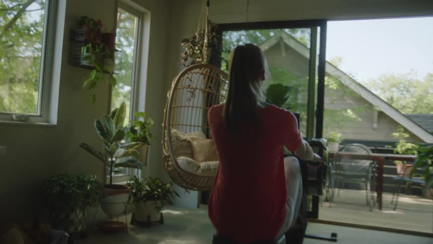 Woman exercises on rowing machine at home near balcony with view of neighbors house