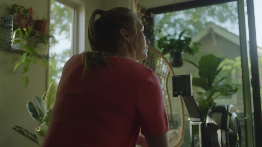 A woman wearing a red shirt stands inside her house looking out the window on a sunny day