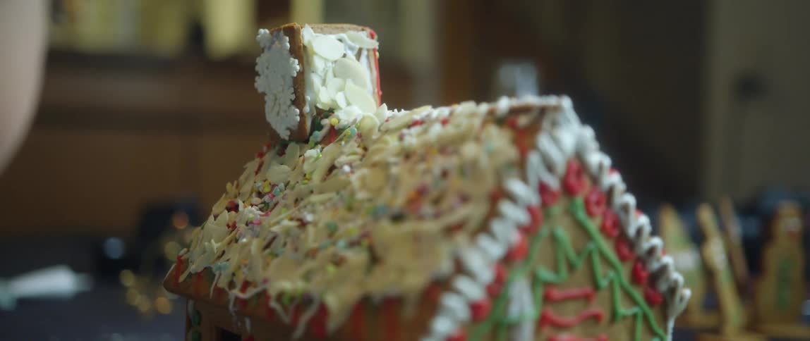 A young girl sits at her dining room table next to a gingerbread house being decorated