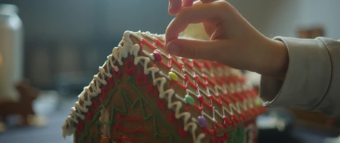 A childs hand places a candy piece on a decorated gingerbread house