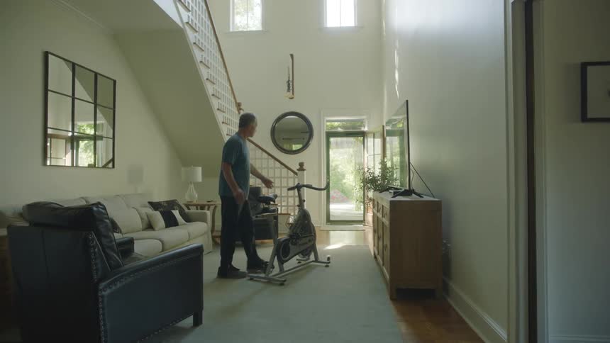 A man walking into his living room and getting on an elliptical