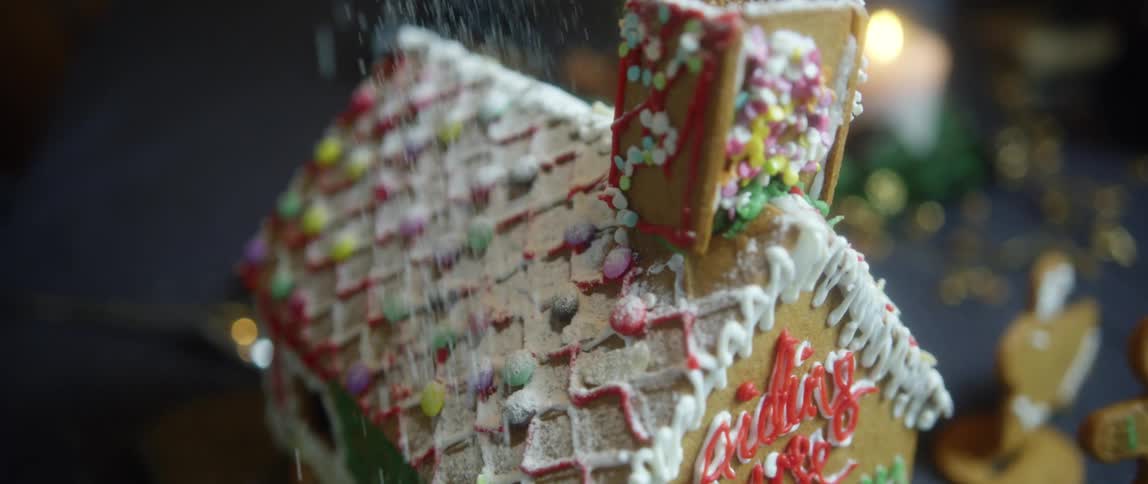 Powdered sugar is being placed on the roof of a gingerbread house decorated with icing sitting on a blue table
