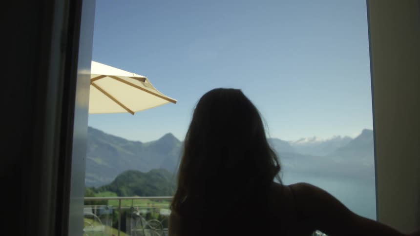A woman pushes drapes and exits onto a balcony overlooking a lake and mountains on a sunny day