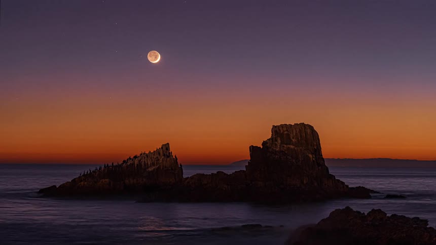The moon moves lower as dusk turns to night over the ocean