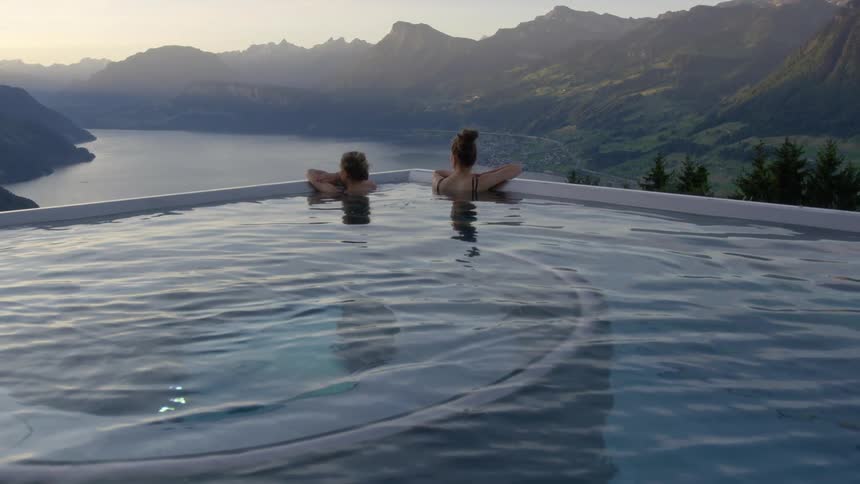 Two women standing on the edge of a pool looking at the lake in the mountains at sunset