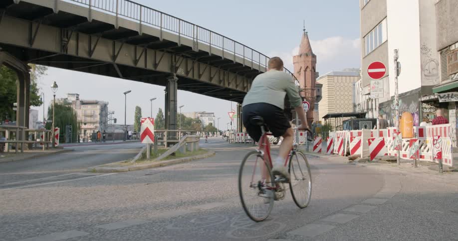 A man rides a bike on a city street with an area of construction and a bridge to his left