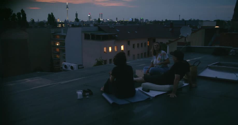 Three people sit on a city building rooftop together talking