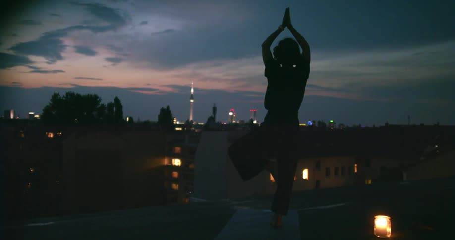 A woman takes a yoga pose while standing on one foot on a yoga mat on the rooftop of a building in a city at sunset