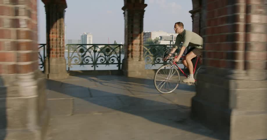 Man bicycles along covered walkway across water from city skyline passing pedestrians