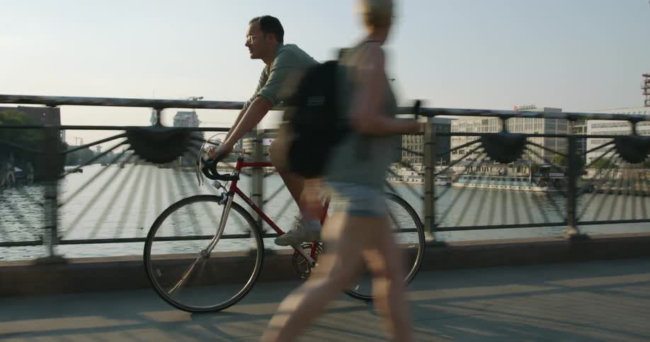 Man pedals bicycle across bridge over river passing others with construction site and city center in background
