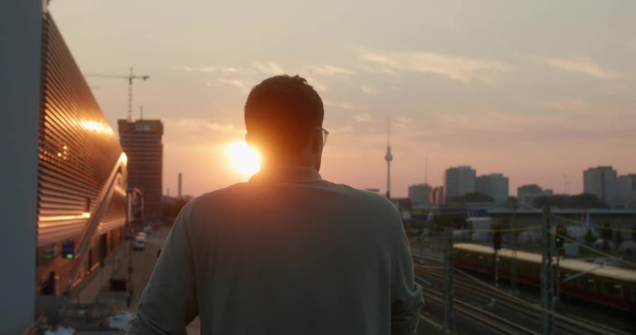 Man standing on an overpass over the train tracks near a train station in the city at sunset